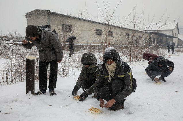 Imigrantes comem o prato de comida que receberam a céu aberto enquanto a neve cai em Belgrado.