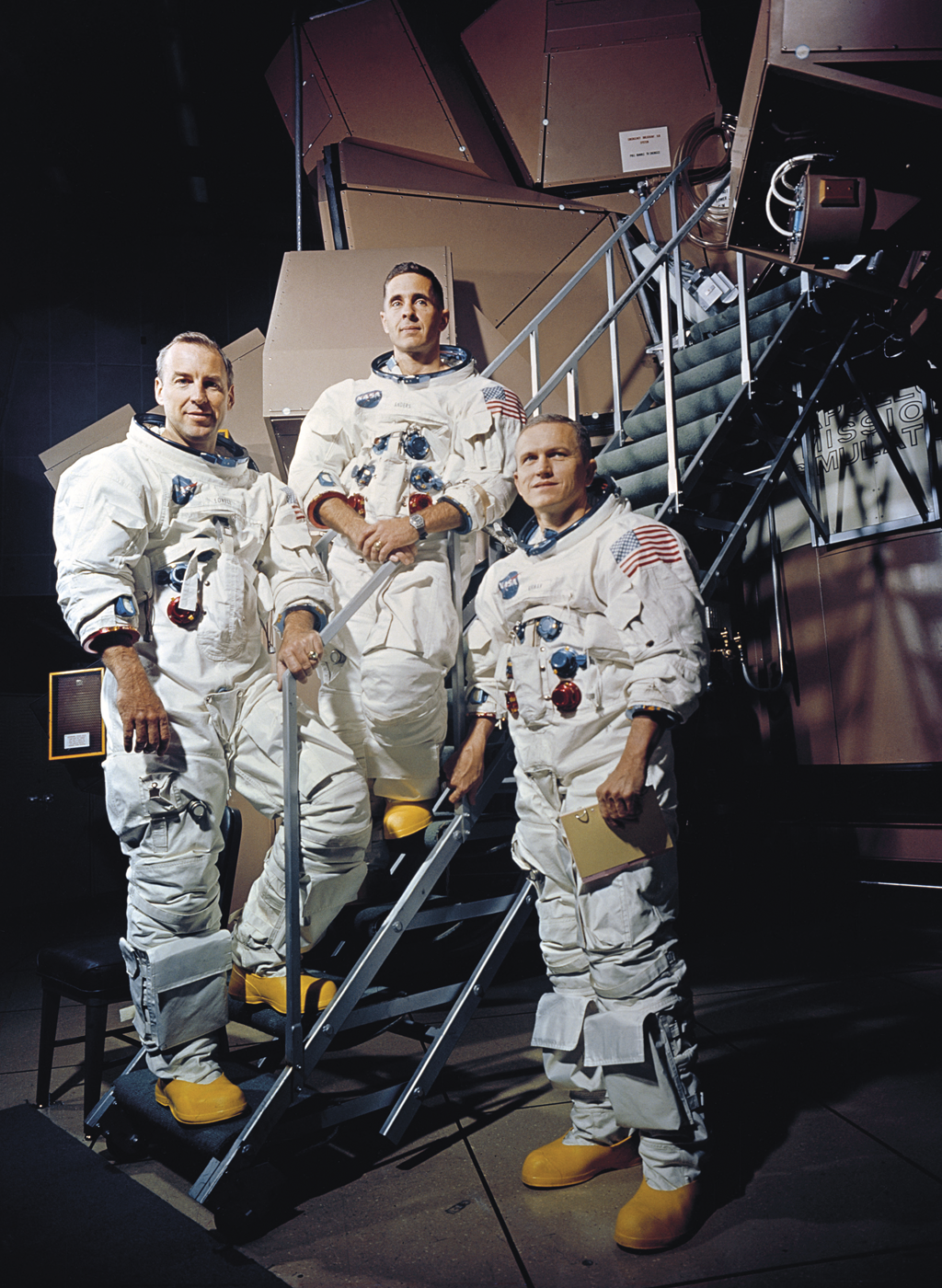 Foto da tripulação da Apollo 8 durante treinamento. Eles estão com os trajes de astronauta no pe de uma escada.