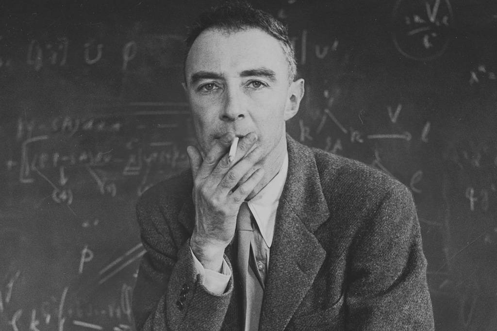 Retrato de Robert Oppenheimer de terno e com um cigarro na boca. No fundo um quadro negro com equações desfocado.