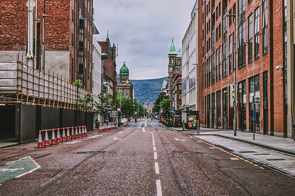 Vista de uma rua vazia no centro de Belfast, na Irlanda, sem carros ou pessoas, com prédios de tijolinho e montanhas ao fundo.