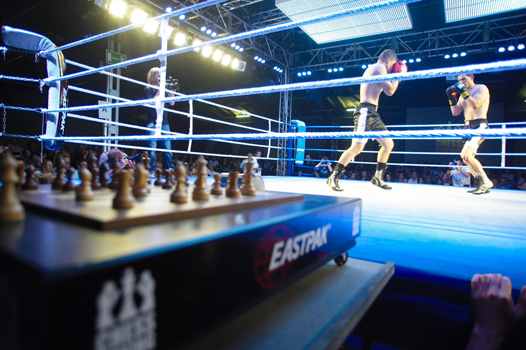 Tabuleiro de xadrez no primeiro plano e, ao fundo, dois homens se enfrentando em um ringue de boxe.