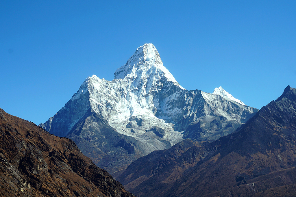 Foto do pico do monte Everest, coberto por neve no topo, e um céu azul sem nuvens por trás.
