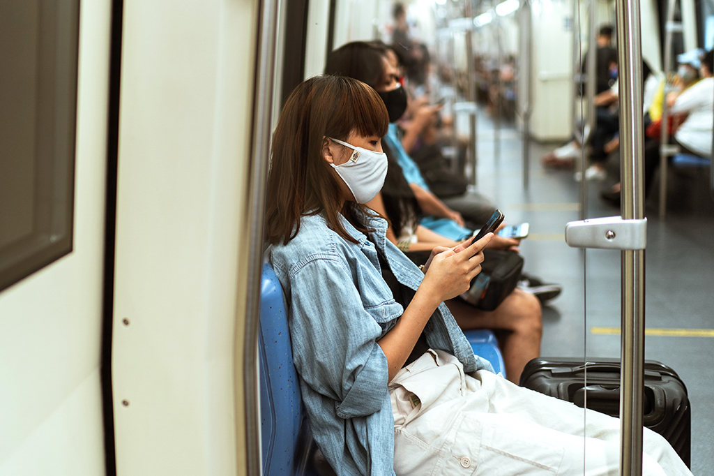 Visão lateral de uma mulher sentada no banco do metrô, de máscara, digitando no celular. Algumas pessoas, também de máscara, ocupam o vagão atrás dela.