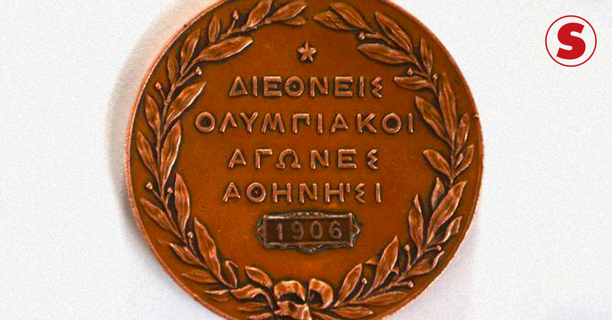 Medalha redonda de bronze, das Olimpíadas de 1906. Ramos de hortelã acompanham a circunferência da medalha e, ao centro, frase em grego com o número "1906".