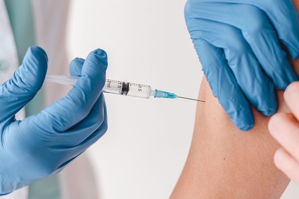 Pessoa com luvas cirúrgicas azuis vacinando o braço direito de outra pessoa.