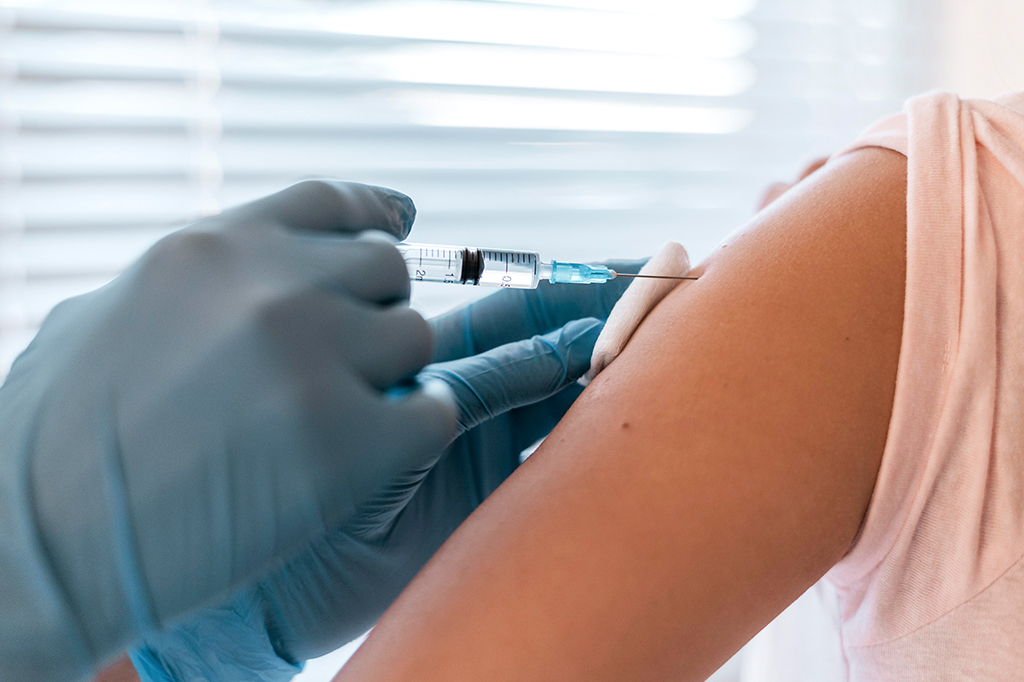 Pessoa com luvas cirúrgicas azuis aplicando uma vacina no braço de outra pessoa.