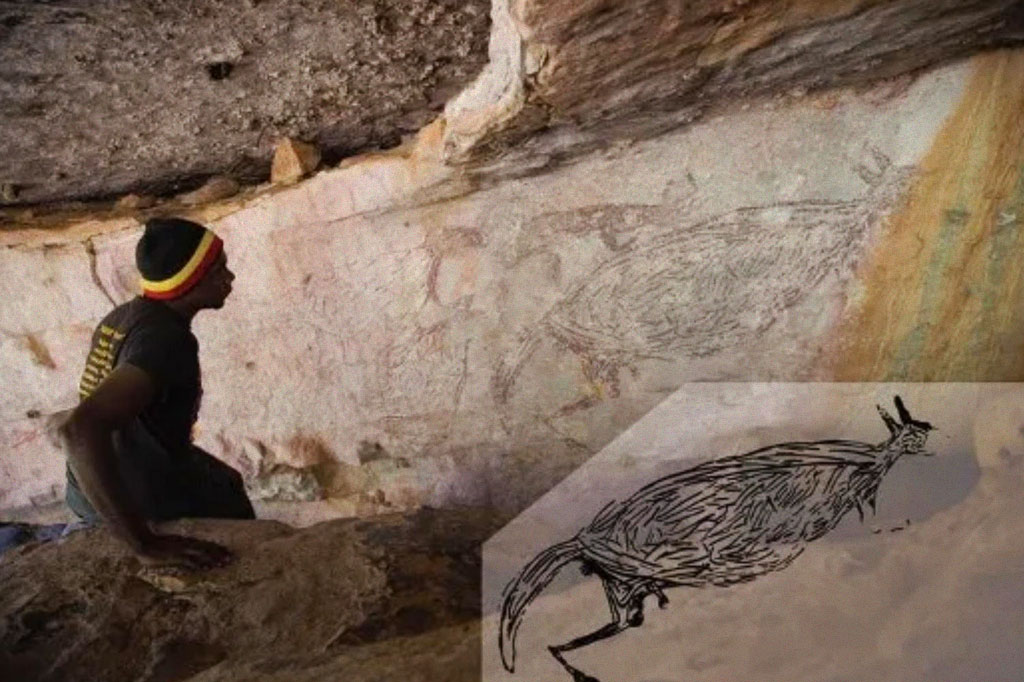 Foto de uma caverna com um homem à esquerda e a pintura rupestre de um canguru à direita.