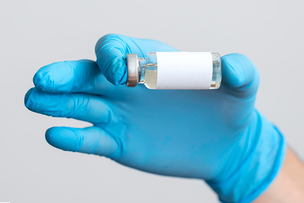 Mão direita de uma pessoa, com luva cirúrgica azul, segurando uma ampola de vacina.