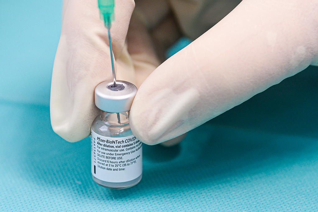 Mão com luva cirúrgica segurando um frasco da vacina Pfizer-BioNTech.