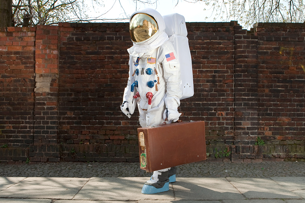 Foto de uma pessoa vestida de astronauta na rua, carregando uma maleta.