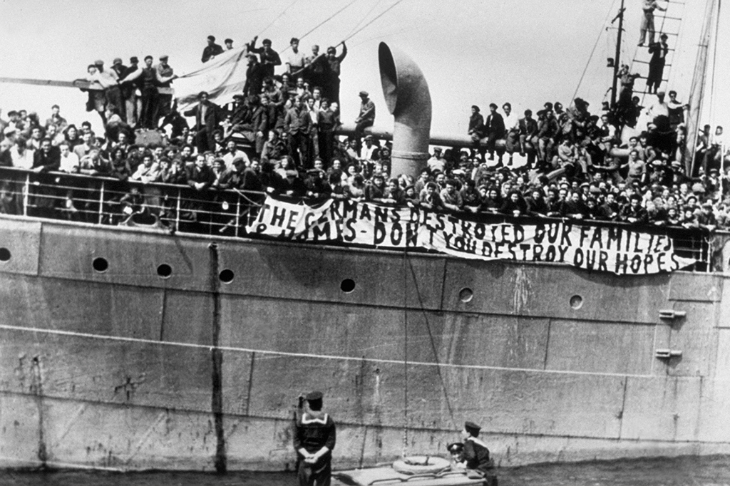 Foto em preto de branco de um navio abarrotado de gente sendo ancorado no porto. São imigrantes judeus ilegais chegando à Palestina em 1947: “Os alemães destruíram nossas famílias, não destruam nossa esperança”, diz a faixa branca colocada no lado do navio.