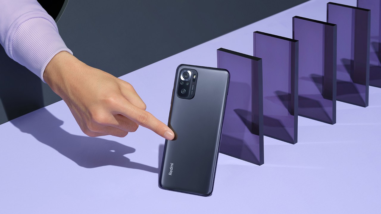 Smartphone Xiaomi Redmi Note 10S na cor preta, num fundo lilás, sendo empurrado por um dedo, com várias placas de acrílico roxas formando uma fila atrás do celular.