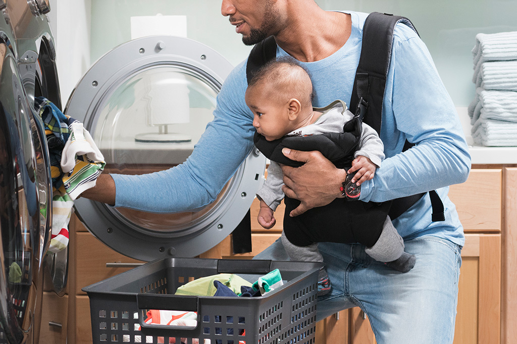 Homem colocando roupas para lavar enquanto seu bebê, preso a sua frente, em um canguru, observa tudo.