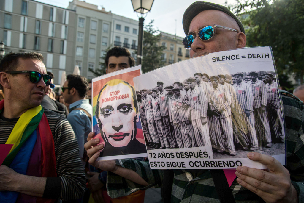 Imagem de homem durante protesto segurando uma placa com o rosto de Vladimir Putting escrito "pare a homofobia" e outra comparando os assassinatos por homofobia com um campo de concentração nazista.