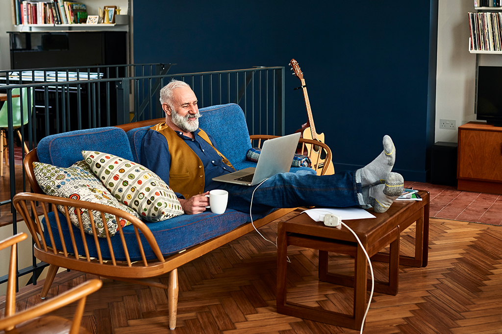 Homem de idade mexendo no computador em sua sala de estar, com coisas ao redor que remetem seus hobbies.