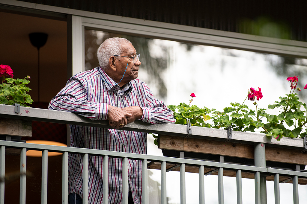 Homem idoso de óculos e camisa listrada apoiado no parapeiro de uma varanda, sozinho, ao lado de um vaso de flores.