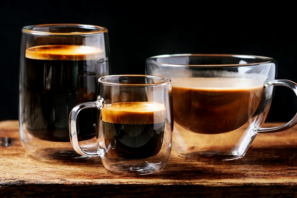 Três xícaras diferentes de vidro com café em cima de uma bandeja de madeira.