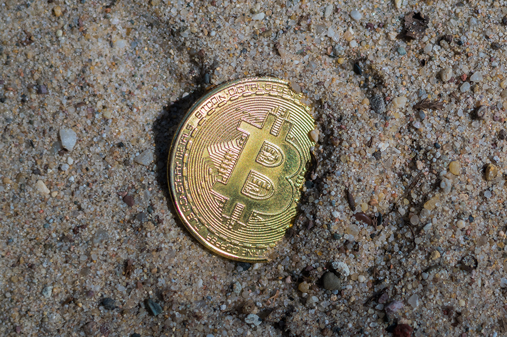 Moeda com símbolo de bitcoin enterrada na areia.
