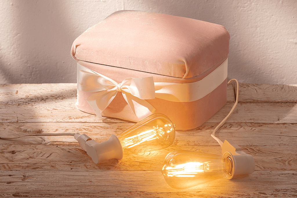 Foto de uma caixa rosa de veludo tampada, com um laço em volta e com duas lâmpadas acesas na frente dela.
