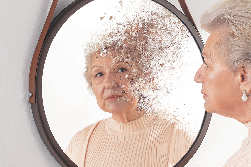 Retrato de uma mulher 60+ se olhando no espelho. Seu rosto no reflexo está rachado e se desfazendo.
