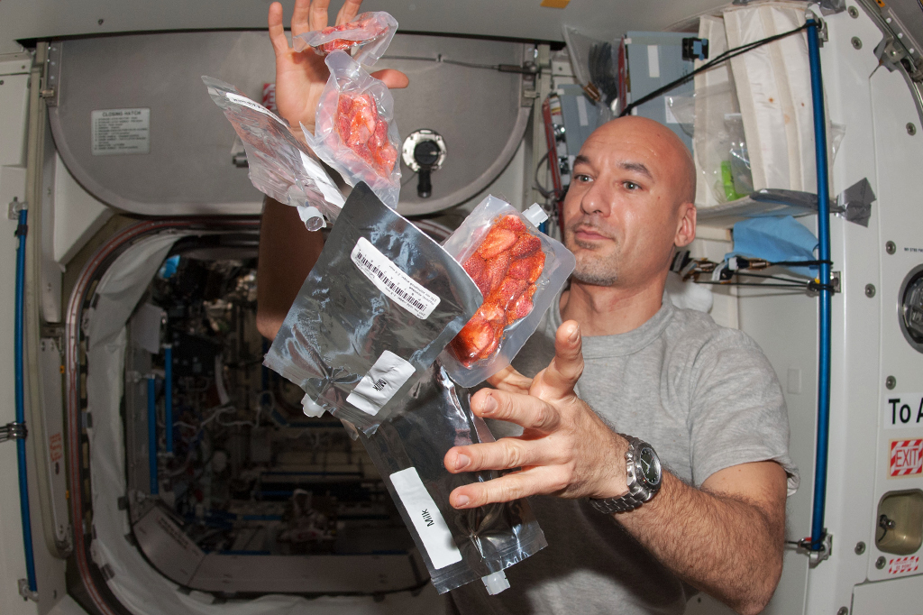 Astronauta em estação espacial com embalagens de alimento flutuando sem gravidade.