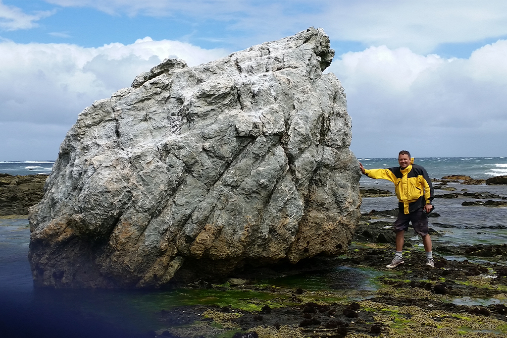 Imagem de um homem, em pé, apoiando a mão direita em uma grande rocha, perto do mar.