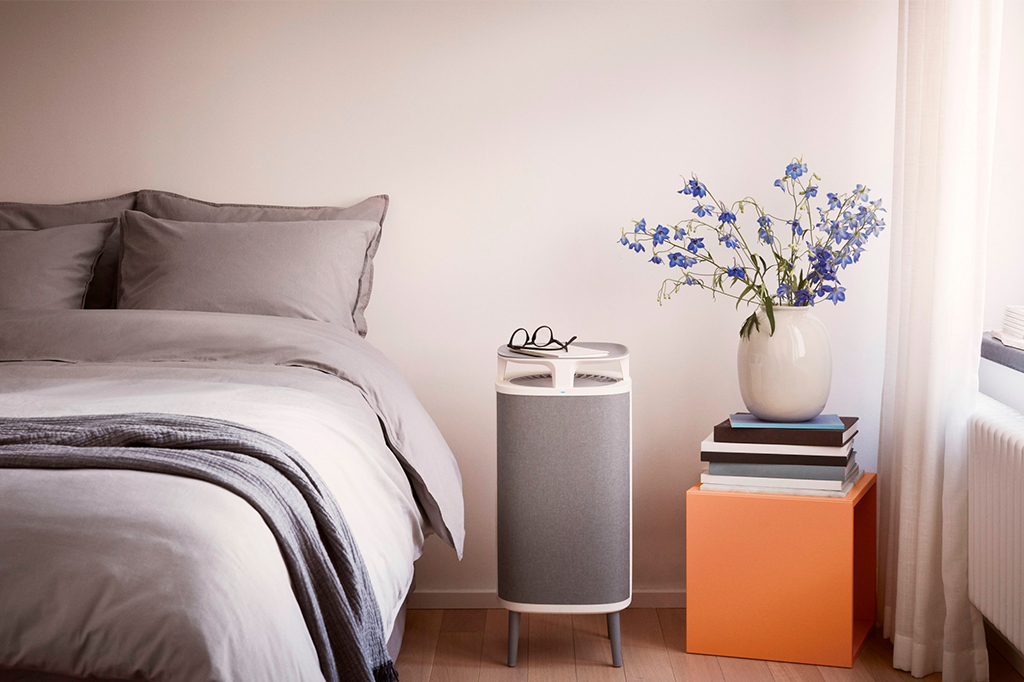 Imagem de um quarto, com detalhe para o filtro de ar que encontra-se entre a cama e um apoio de cabeceira com livros e vaso de flor.