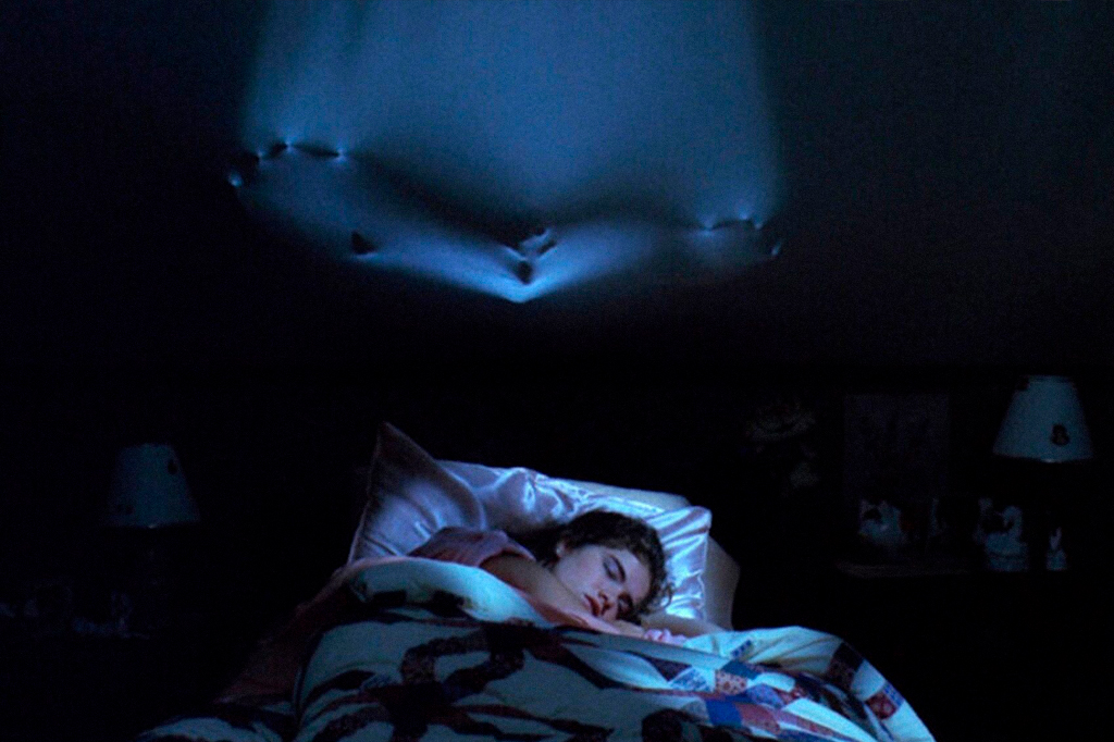 Cena de 'A hora do pesadelo', em que uma personagem está dormindo na cama. Acima dela, na parede do quarto, uma figura estranha parece assombrá-la.