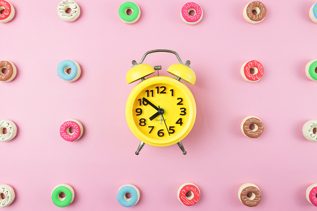 Fotografia de um relógio amarelo em cima de uma mesa com donuts ao redor dele.