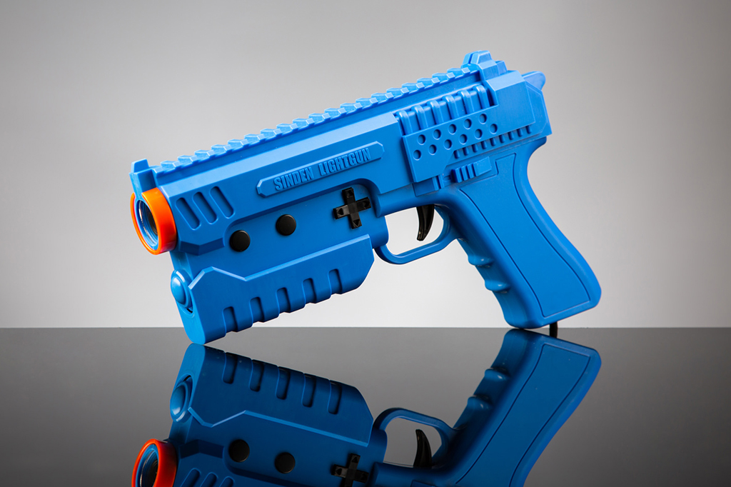 Fotografia da pistola Sinden Lightgun, que é de cor azul com detalhe em laranja, apoiada em superfície cinza espelhada.