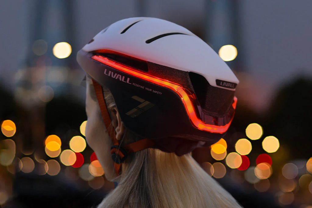 Fotografia noturna de uma mulher de costas, vestindo o capacete Livall, que está com as luzes acesas. No fundo, vê-se pontos de luz desfocados da rua.