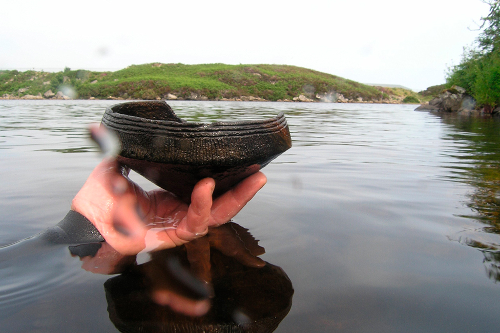 Fotografia de uma mão segurando um pote de cerâmica, molhado, em um lago.