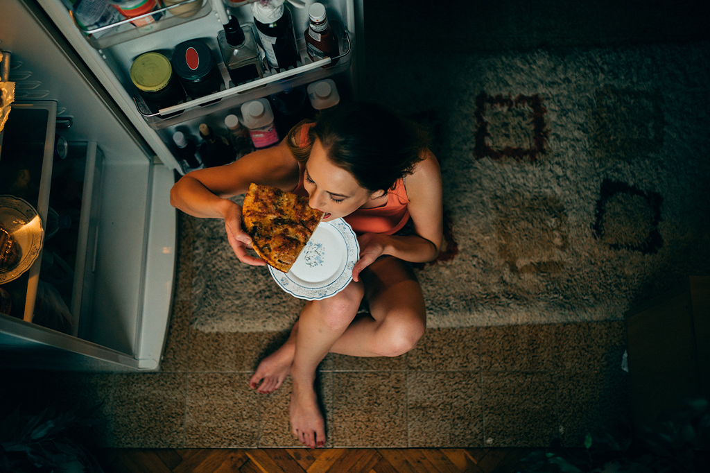 Mulher sentada num tapete em frente a uma geladeira aberta, à noite, comendo um pedaço de pizza.