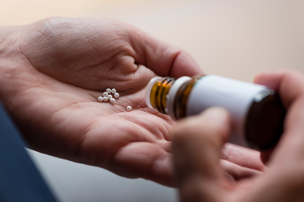 Uma pessoa derramando pílulas homeopáticas (glóbulos) de um frasco de vidro na palma da mão.