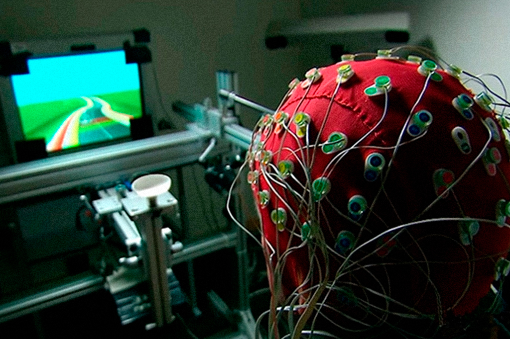 Pessoa vestindo touca com diversos fios analisando a atividade cerebral enquanto joga videogame em laboratório.