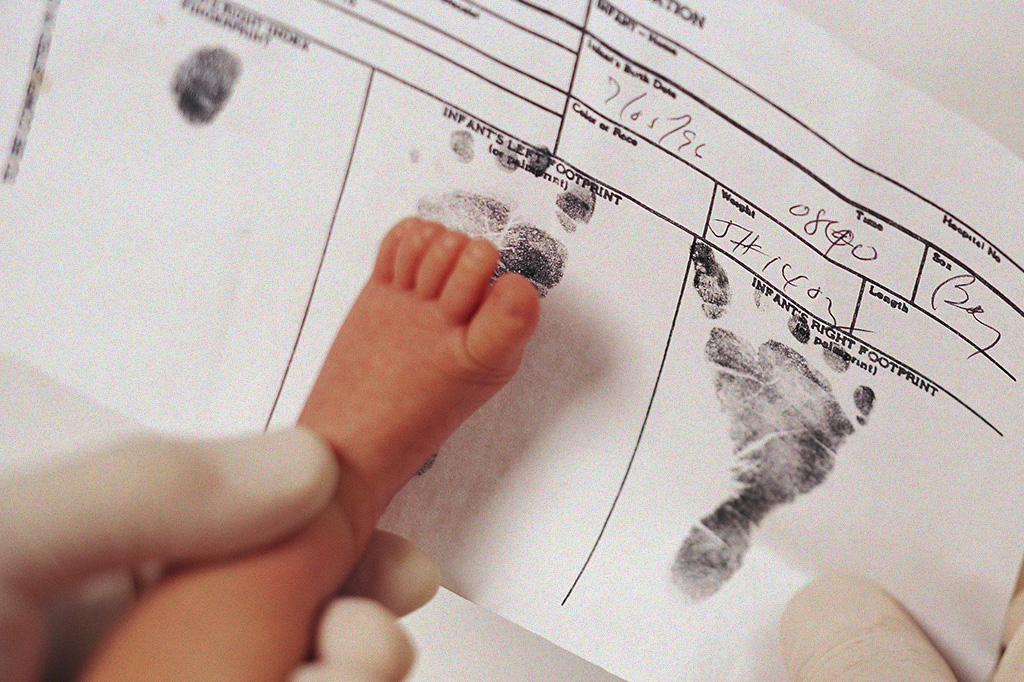 Foto mostrando as mãos de um médico carimbando um pézinho de bebê na certidão de nascimento.