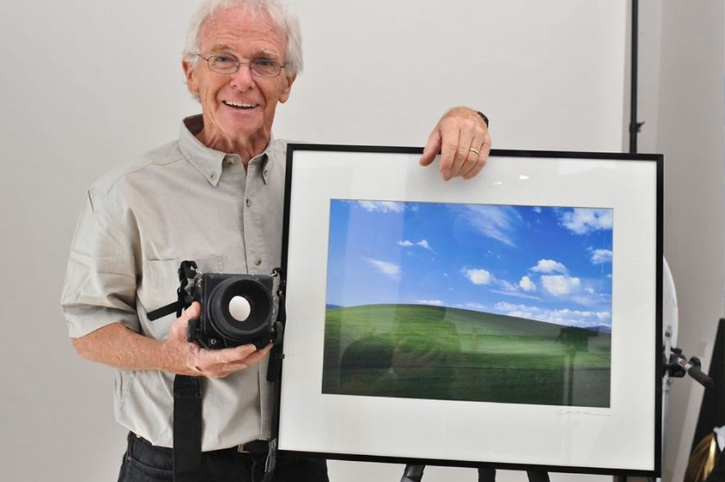 Retrato do fotógrafo Charles O'Rear ao lado de um quadro com a fotografia de 'Bliss' emoldurada.