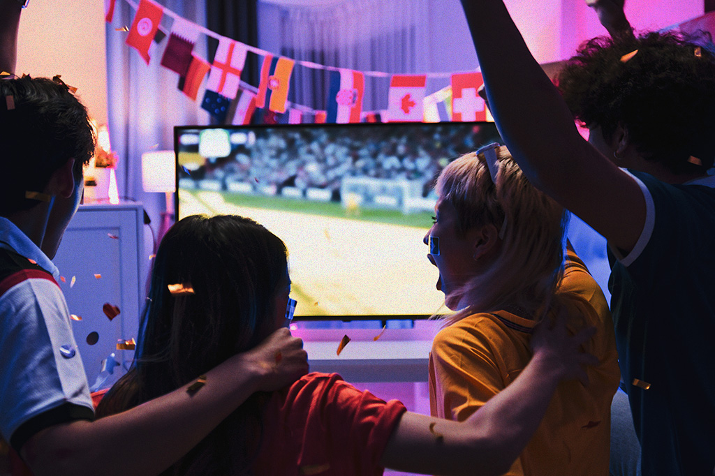 Grupo de jovens felizes assistindo a um jogo de futebol na televisão de casa e comemorando um gol juntos.