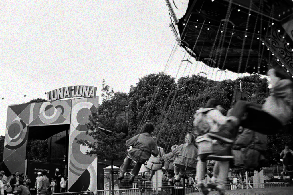 Foto em preto e branco na qual podemos ver o letreiro do parque Luna Luna e algumas crianças brincando no brinquedo 