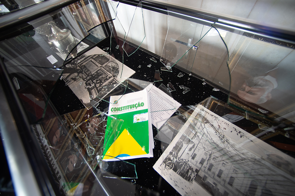 Foto de cacos de vidro quebrados em cima de imagens e do livro da Constituição no Museu do Senado.