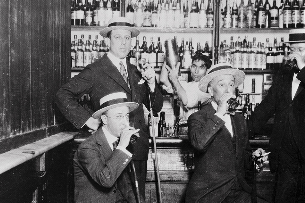 Homens em um speakeasy bebericando drinks. Ao fundo, um barman prepara uma bebida com uma coqueteleira.