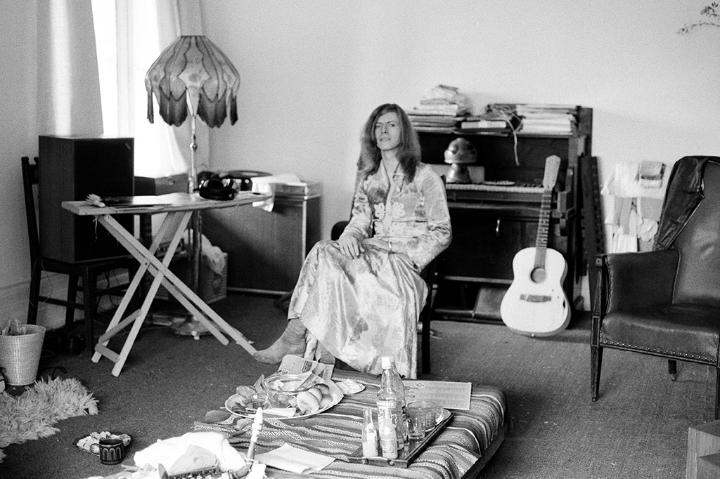 Retrato preto e branco de David Bowie em sua casa, vestindo o vestido e sentado em uma cadeira em um cômodo com violão, piano, poltronas e louças de vidro.