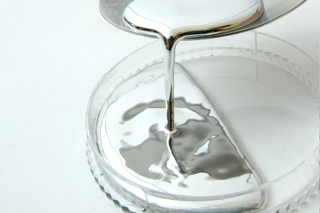 Mercúrio líquido sendo despejado com uma espátula em um recipiente de plástico transparente.