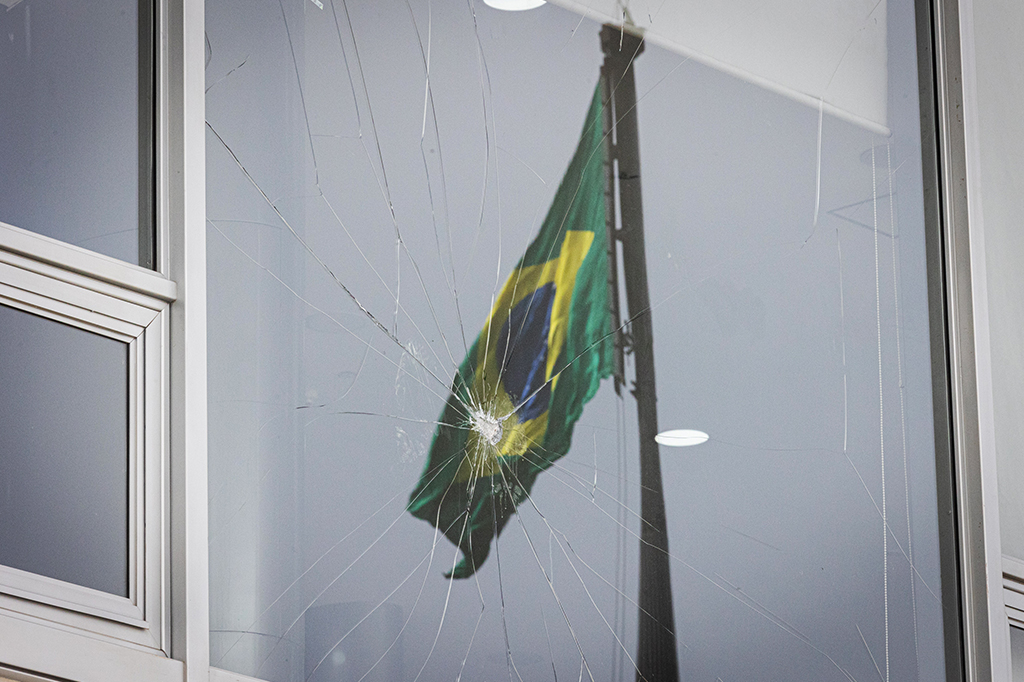 Foto da bandeira do Brasil refletida na janela quebrada do Palácio do Planalto.