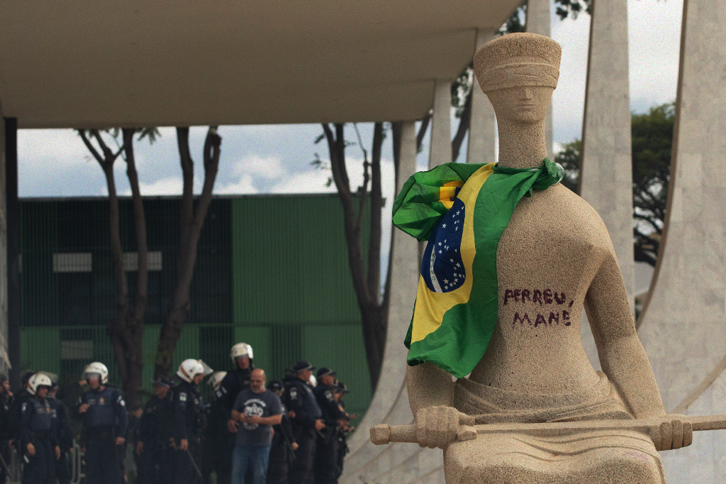 Foto da escultura "A Justiça" pixada e com a bandeira do brasil amarrada no pescoço.