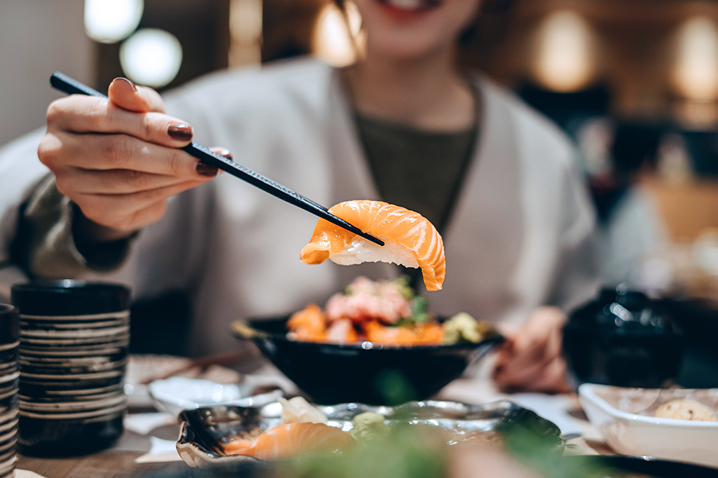Uma mulher pegando uma peça de sushi com hashis, em primeiro plano. Ao fundo, desfocado, vê-se a silhueta da mulher, que está sentada à mesa.