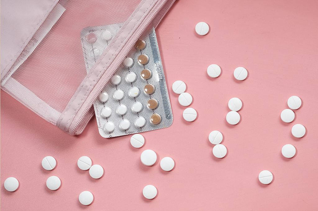Cartela de pílula contraceptiva e comprimidos espalhados em fundo rosinha claro.