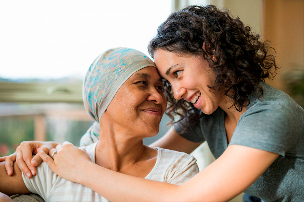 Foto de mulher negra adulta paciente de câncer abraçando sua filha. Ambas sorriem.