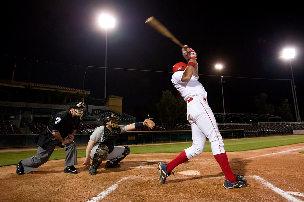 Fotografia de um jogo de baseball, com o rebatedor segurando um taco, em movimento.