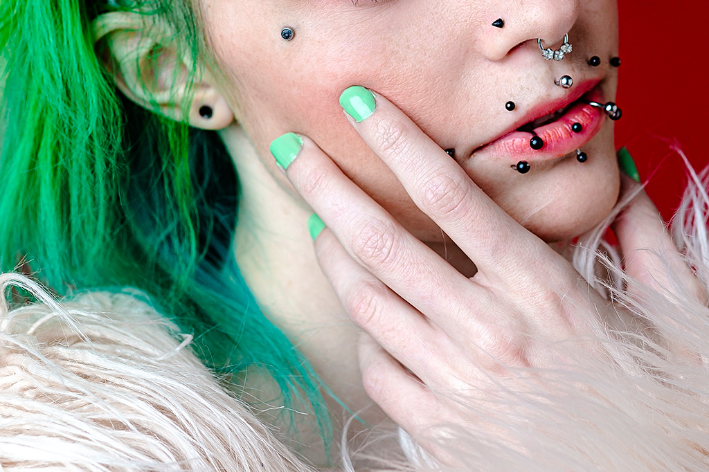 Foto de uma pessoa branca, de cabelo verde, com vários piercings na boca, nariz e rosto.